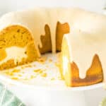 A pumpkin bundt cake with cream cheese filling and white icing sits on a cake stand with one slice removed.