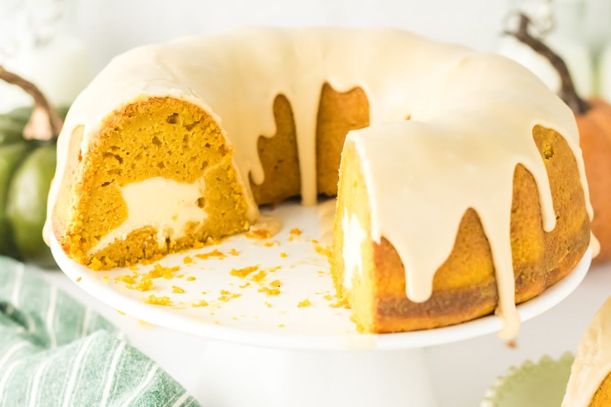 A pumpkin bundt cake with cream cheese filling and white icing sits on a cake stand with one slice removed.