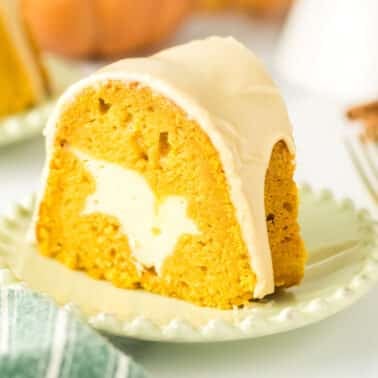 A slice of pumpkin bundt cake with a cream cheese filling and white icing, served on a light green plate with a fork and napkin nearby.