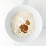 A white bowl containing flour, cinnamon, baking soda, and baking powder sits on a white marble surface.