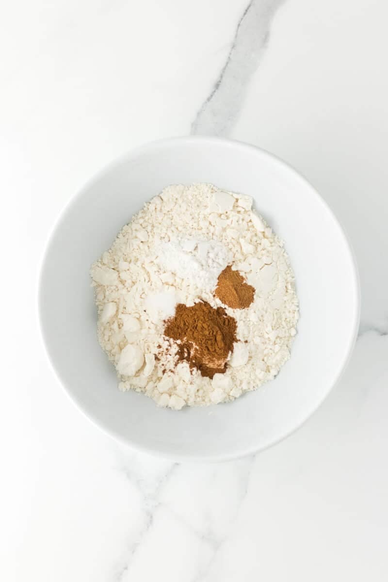 A white bowl containing flour, cinnamon, baking soda, and baking powder sits on a white marble surface.