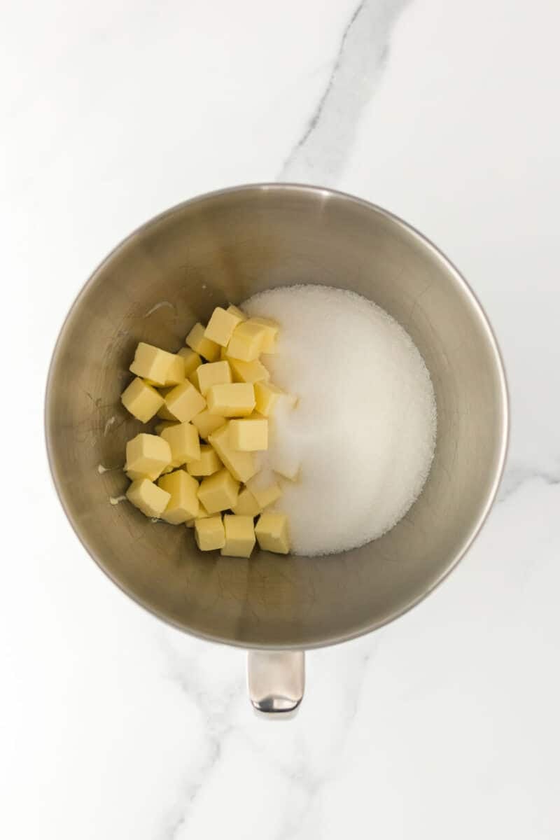 A metal mixing bowl containing cubed butter and granulated sugar on a white marble surface.