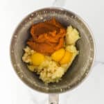 Mixing bowl containing pumpkin puree, eggs, and mashed potatoes on a white marble surface.