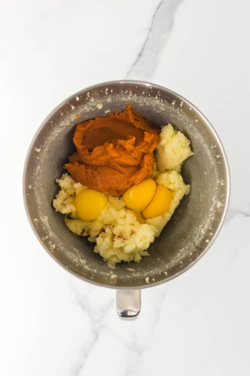 Mixing bowl containing pumpkin puree, eggs, and mashed potatoes on a white marble surface.