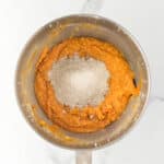 A metal mixing bowl containing orange batter with a mound of flour on top, placed on a white marble surface.