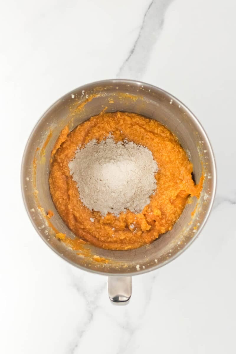 A metal mixing bowl containing orange batter with a mound of flour on top, placed on a white marble surface.