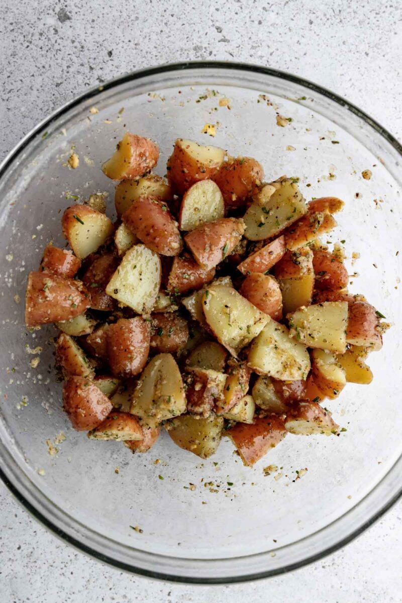 A glass bowl filled with quartered red potatoes coated in herbs and seasoning sits on a light-colored surface.