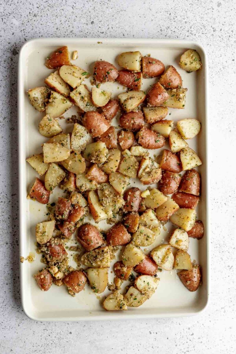 Diced red potatoes coated with herbs and seasoning, spread evenly on a rectangular baking sheet, ready for roasting.