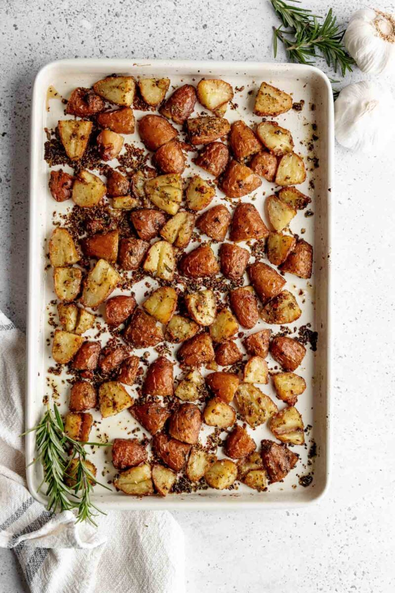 A baking sheet filled with roasted potato pieces garnished with herbs, shown on a light countertop with rosemary sprigs and garlic cloves nearby.