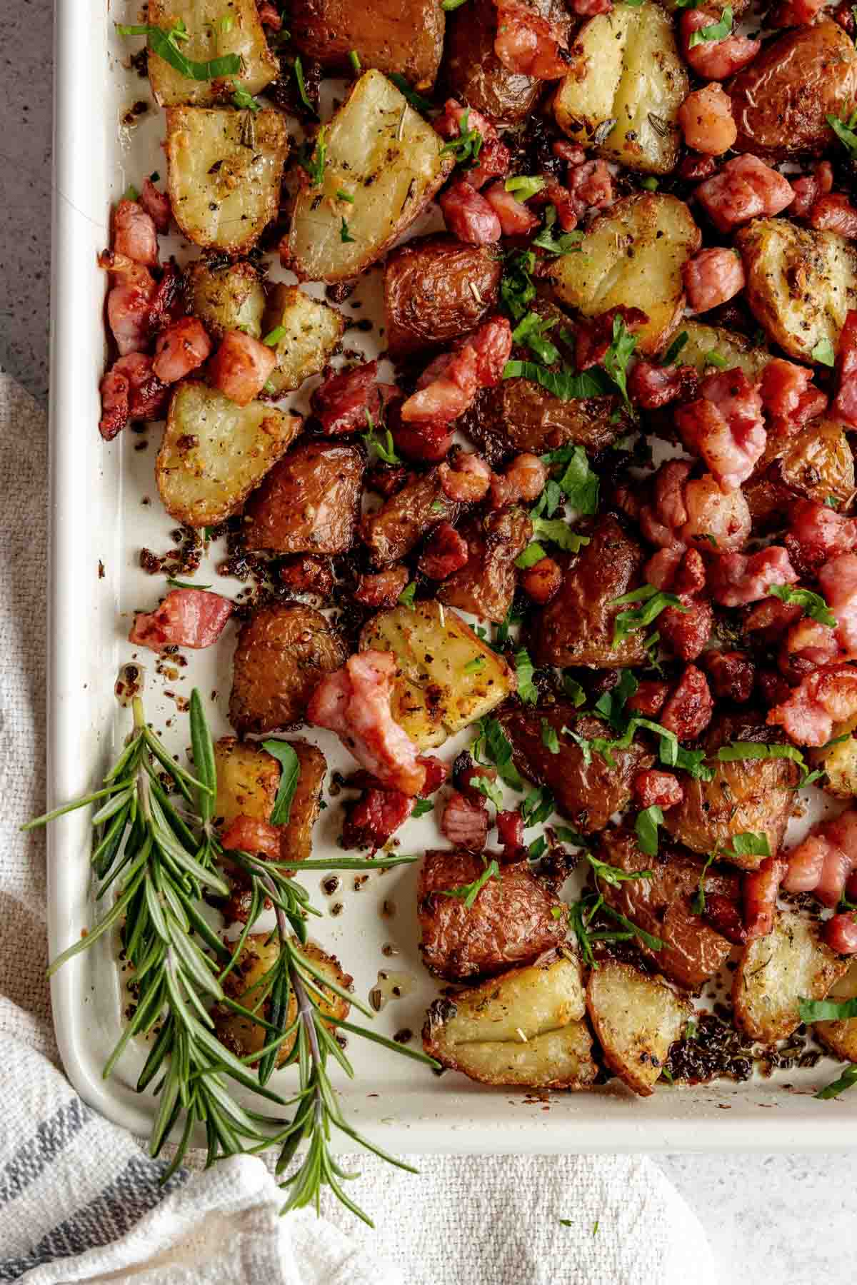 Roasted potatoes and diced bacon pieces garnished with fresh chopped herbs on a white baking tray, with a sprig of rosemary on the side.
