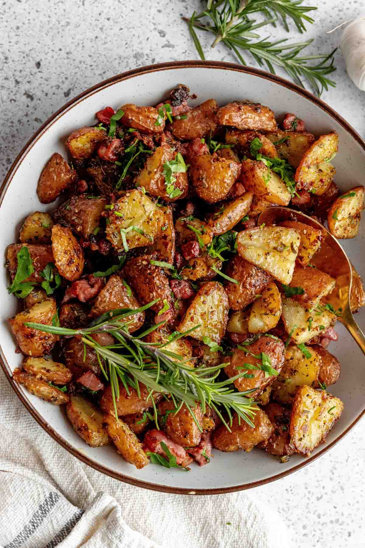 A bowl of roasted potatoes garnished with fresh rosemary and parsley, with visible crispy edges and small bits of bacon.
