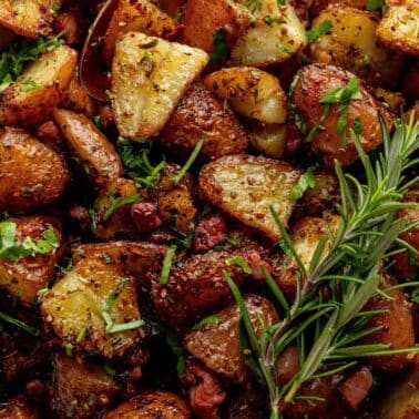 Roasted potato wedges with herbs and bacon pieces, garnished with fresh parsley and a sprig of rosemary, served in a white bowl.