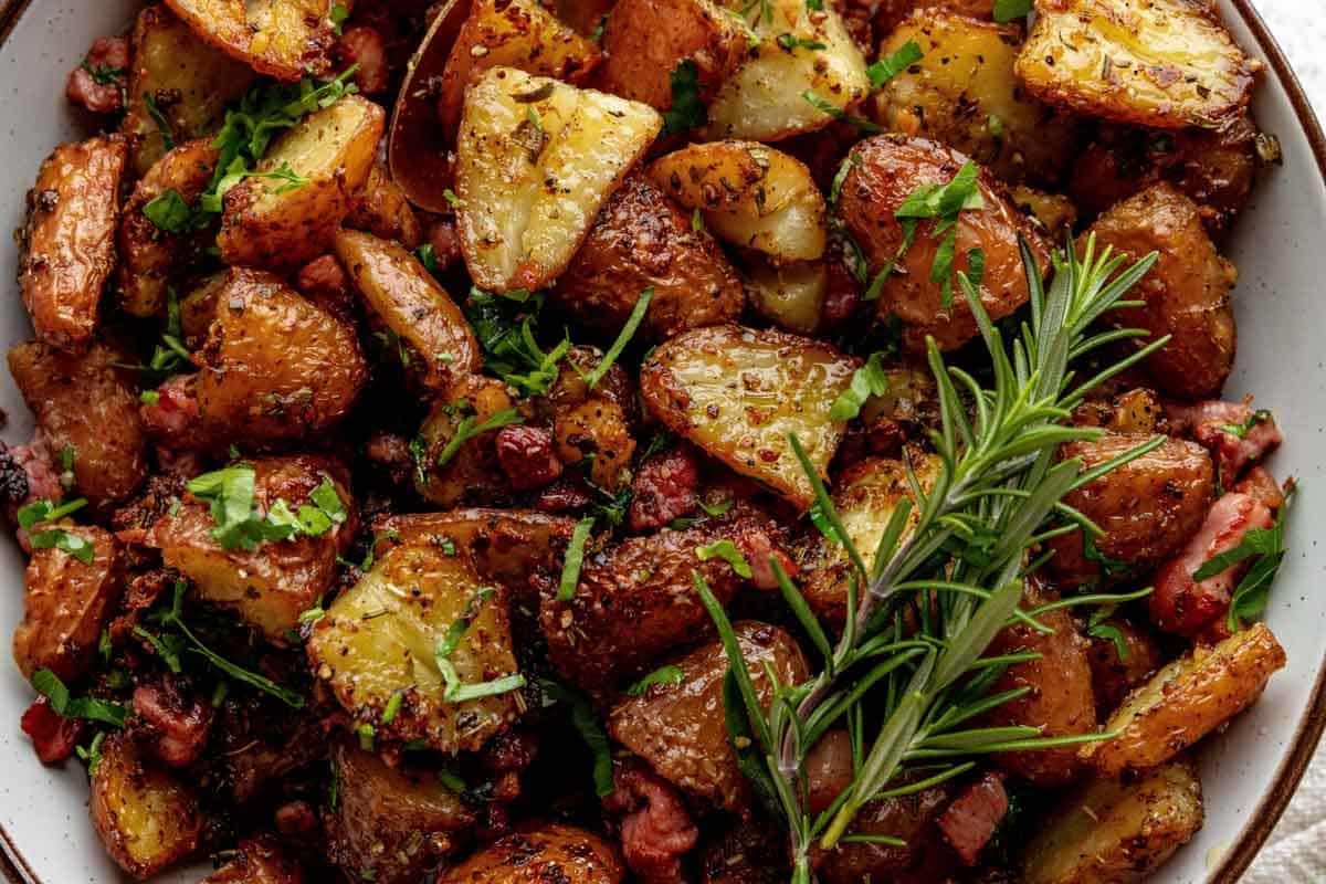 Roasted potato wedges with herbs and bacon pieces, garnished with fresh parsley and a sprig of rosemary, served in a white bowl.
