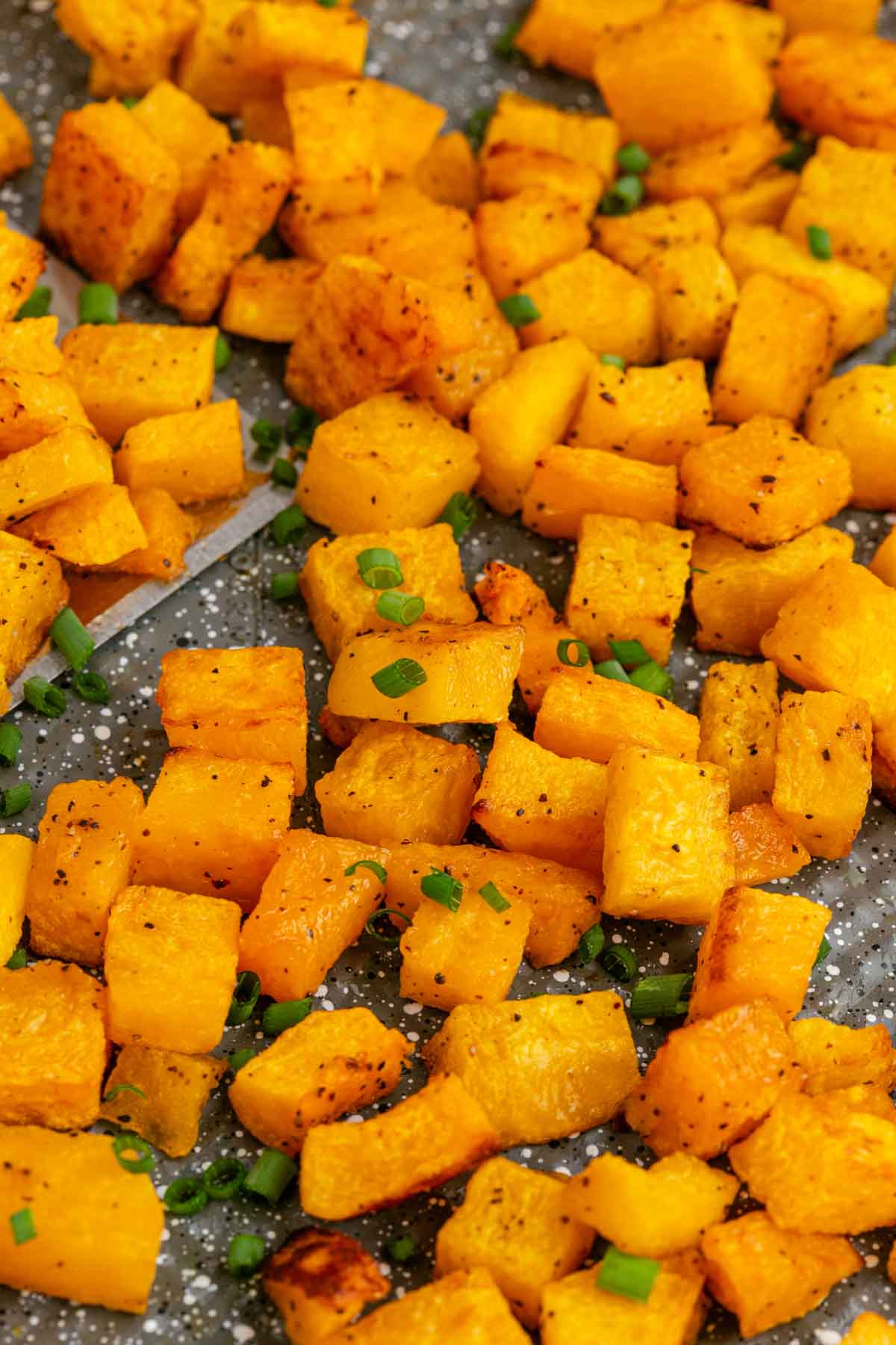 Cubed roasted butternut squash pieces seasoned with black pepper and garnished with chopped green onions on a speckled baking sheet.