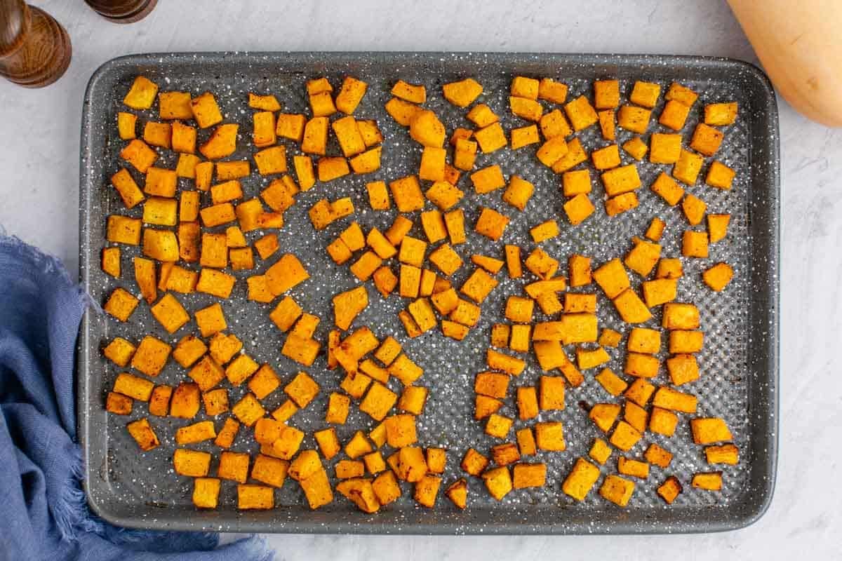 Roasted butternut squash cubes are spread evenly on a baking sheet, seasoned, and baked to a golden brown on a light-colored countertop.