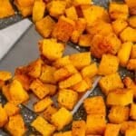 Diced, roasted butternut squash pieces are spread on a baking tray, with a metal spatula lifting some of the seasoned cubes.