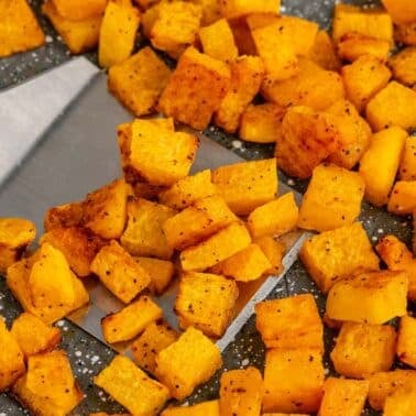 Diced, roasted butternut squash pieces are spread on a baking tray, with a metal spatula lifting some of the seasoned cubes.