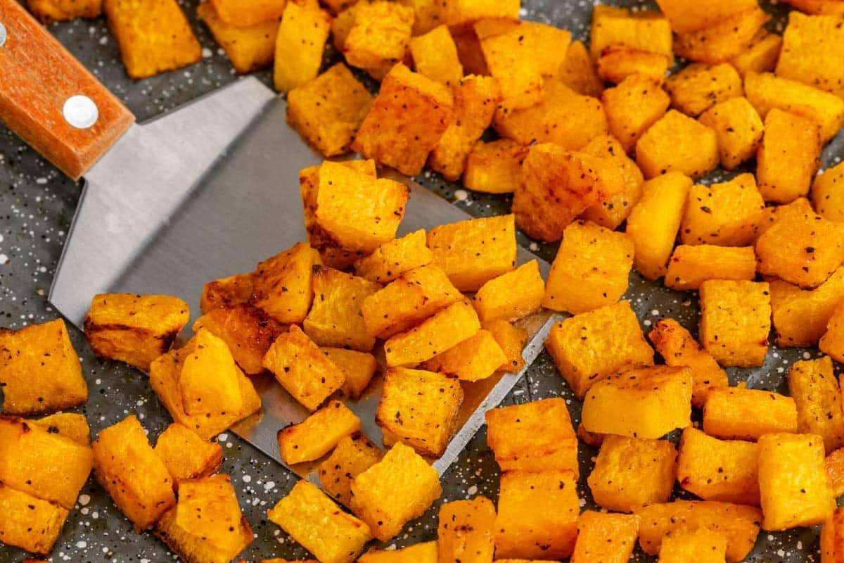 Diced, roasted butternut squash pieces are spread on a baking tray, with a metal spatula lifting some of the seasoned cubes.