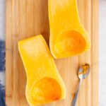 Two halves of a butternut squash with seeds removed, placed on a wooden cutting board next to a metal spoon.