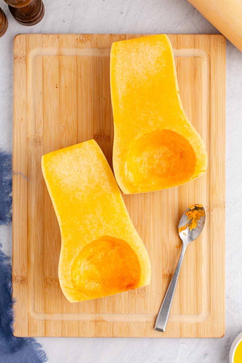 Two halves of a butternut squash with seeds removed, placed on a wooden cutting board next to a metal spoon.