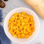 A white bowl filled with cubed butternut squash sits on a light surface near a whole squash, salt and pepper shakers, a blue cloth, and a small dish of oil.