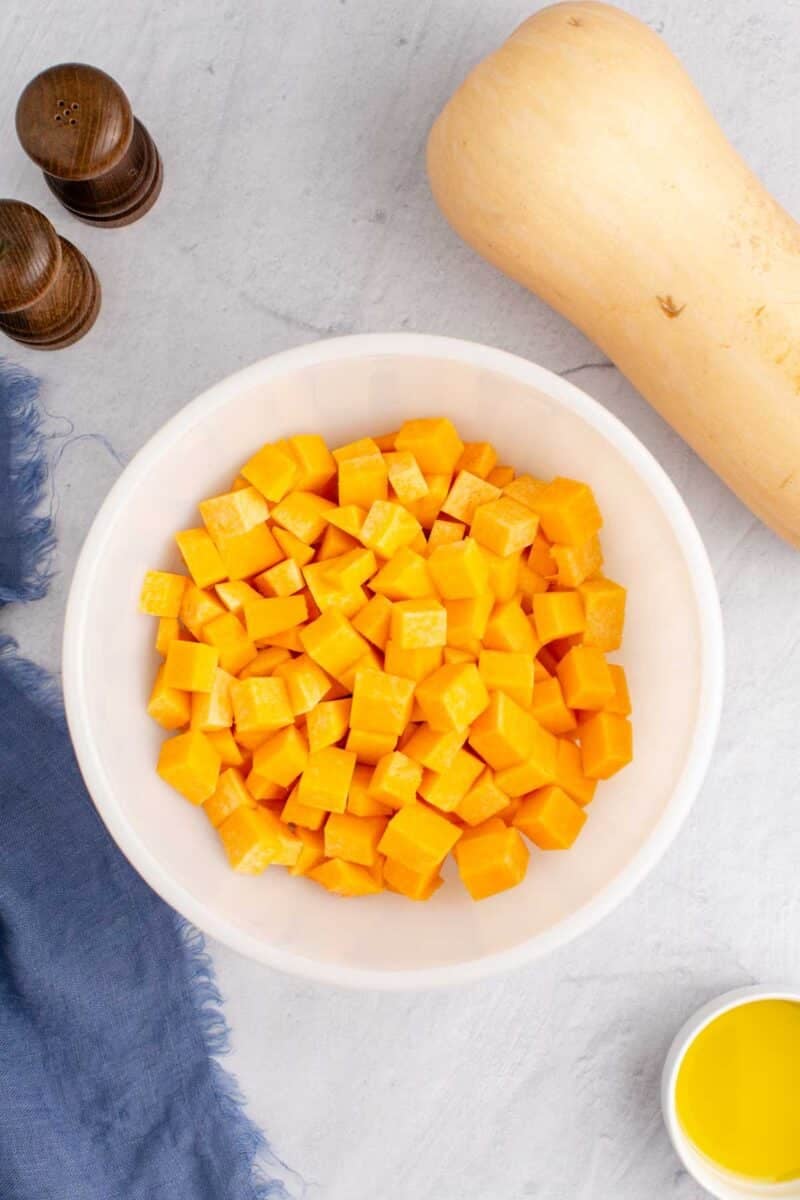 A white bowl filled with cubed butternut squash sits on a light surface near a whole squash, salt and pepper shakers, a blue cloth, and a small dish of oil.