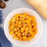 A white bowl filled with cubed butternut squash seasoned with pepper, next to a whole butternut squash, salt and pepper shakers, and a blue cloth on a light surface.