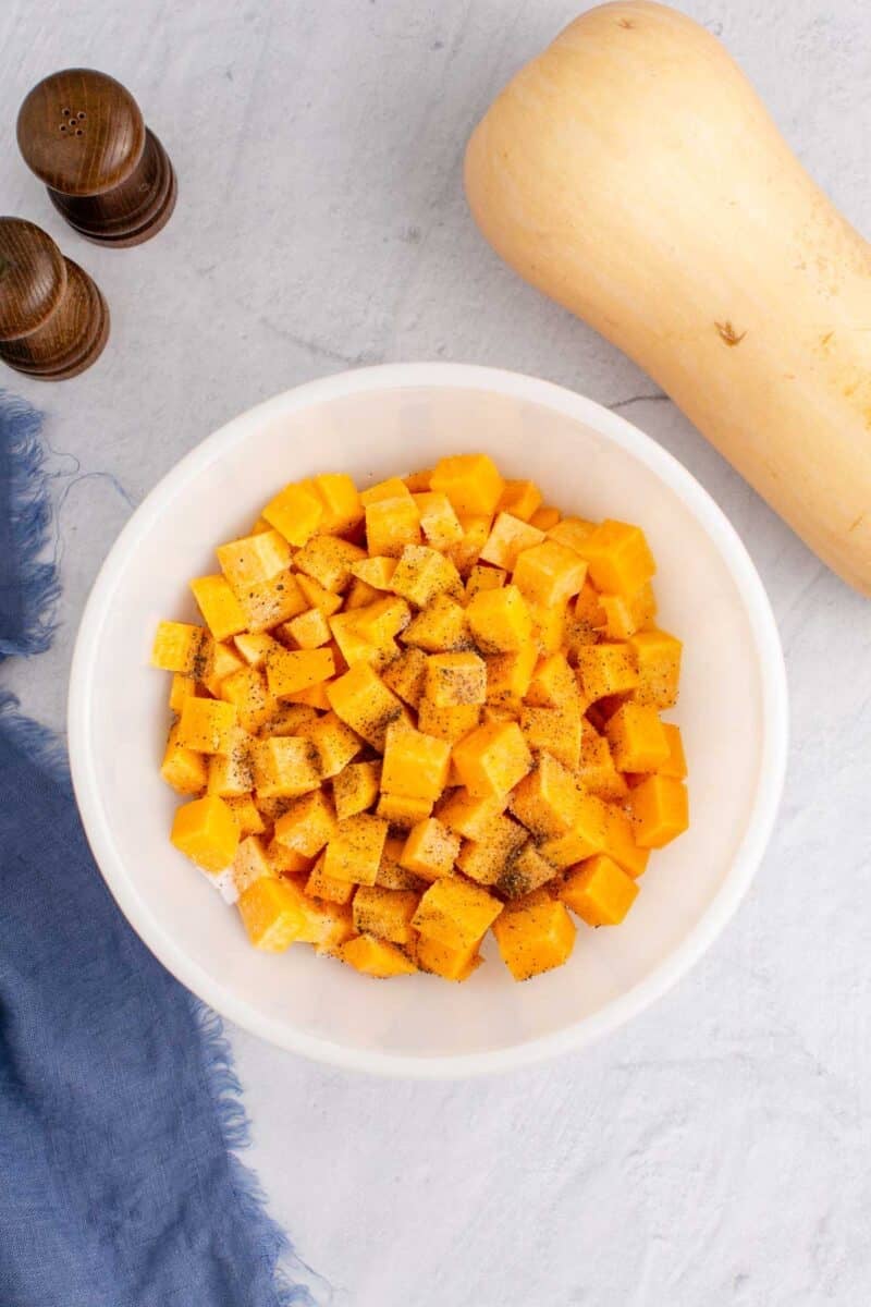 A white bowl filled with cubed butternut squash seasoned with pepper, next to a whole butternut squash, salt and pepper shakers, and a blue cloth on a light surface.