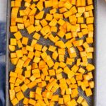 A baking sheet filled with evenly spread, diced butternut squash pieces seasoned with salt and pepper, ready for roasting.