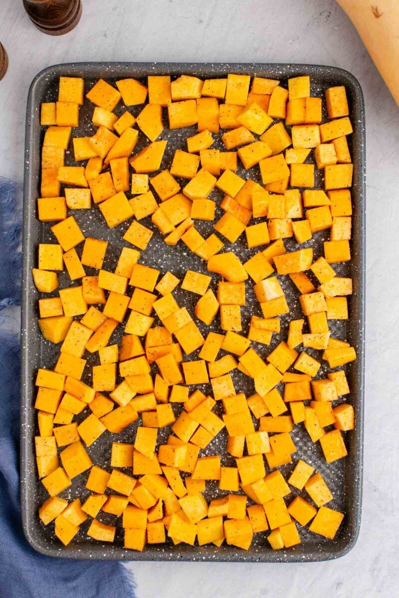A baking sheet filled with evenly spread, diced butternut squash pieces seasoned with salt and pepper, ready for roasting.