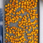 A baking sheet filled with evenly spread, roasted butternut squash cubes on a light gray surface with a blue cloth nearby.