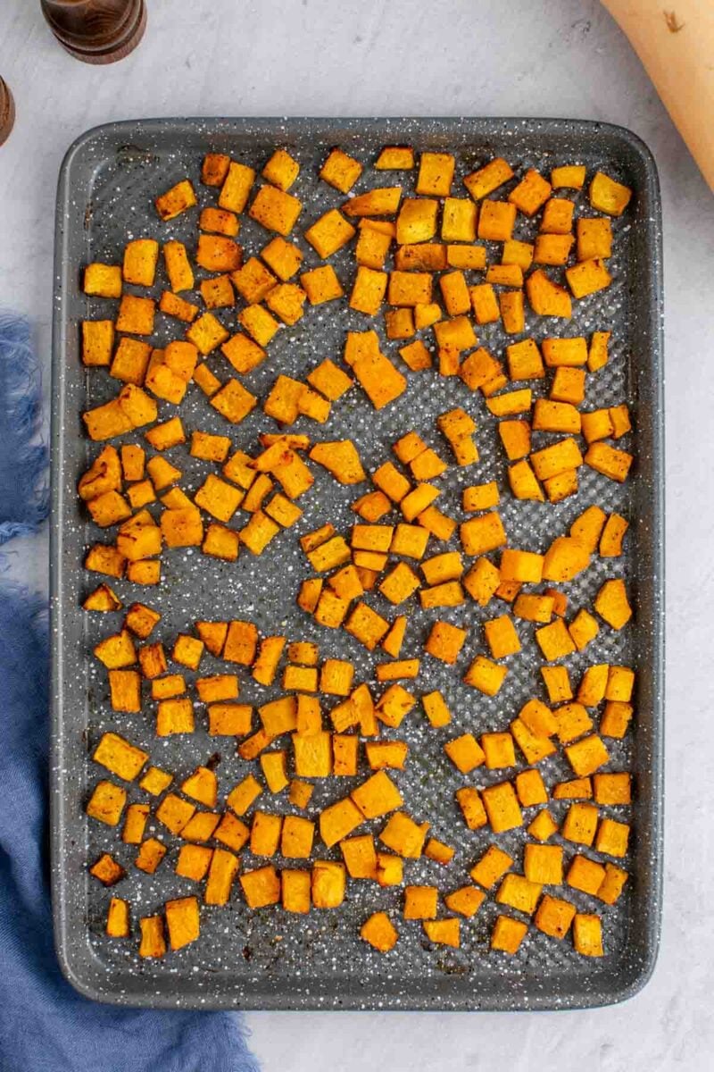 A baking sheet filled with evenly spread, roasted butternut squash cubes on a light gray surface with a blue cloth nearby.