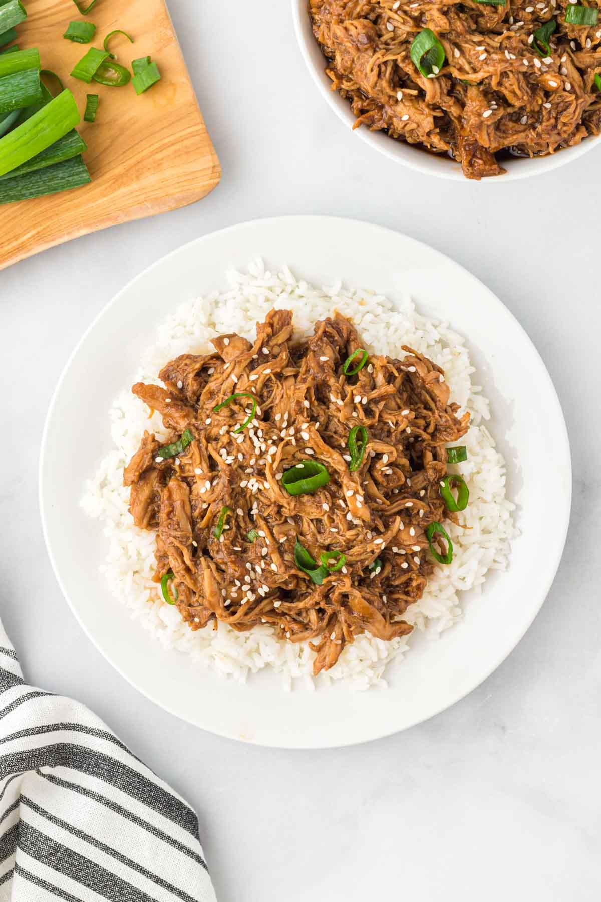A white plate with slow cooker honey garlic chicken in a brown sauce served over white rice, garnished with sesame seeds and green onions. A cutting board with chopped green onions is nearby.