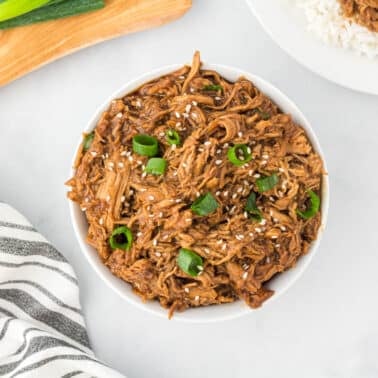 Bowl of shredded meat topped with sesame seeds and chopped green onions, placed on a white surface next to a striped towel and cutting board.