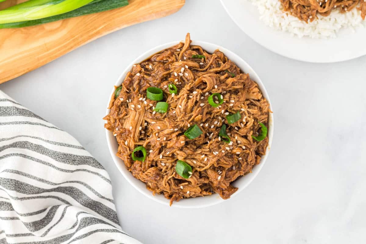 Bowl of shredded meat topped with sesame seeds and chopped green onions, placed on a white surface next to a striped towel and cutting board.