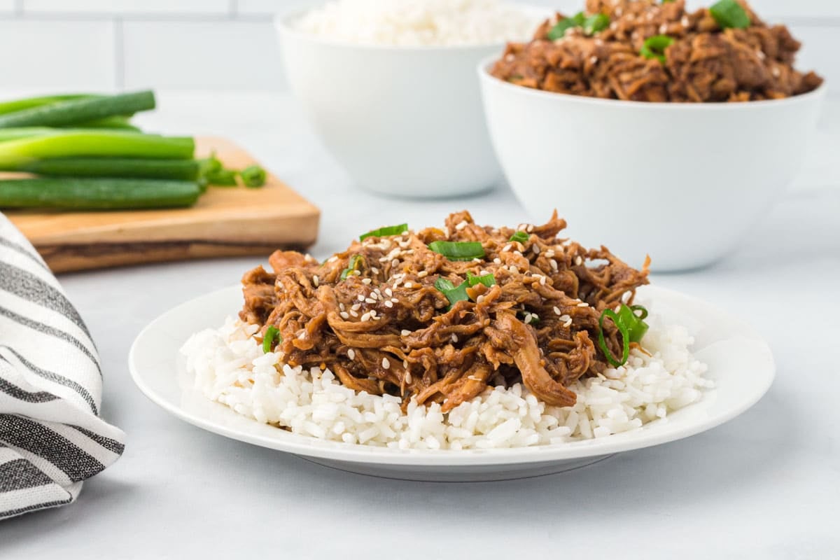 A plate of shredded meat with sauce served over white rice, garnished with sesame seeds and green onions. A bowl of rice and a bowl of meat are in the background.