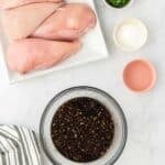 Raw chicken breasts on a plate, a bowl of marinade, chopped green onions, cornstarch, and a small bowl with liquid are arranged on a white surface with a striped cloth nearby.