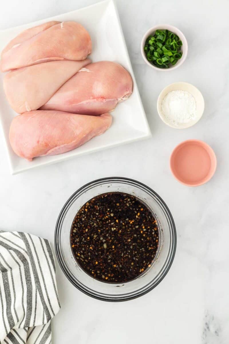 Raw chicken breasts on a plate, a bowl of marinade, chopped green onions, cornstarch, and a small bowl with liquid are arranged on a white surface with a striped cloth nearby.