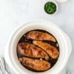 Chicken breasts in dark sauce inside a white slow cooker, with small bowls of sliced green onions, cornstarch, and a pink bowl nearby on a marble surface.