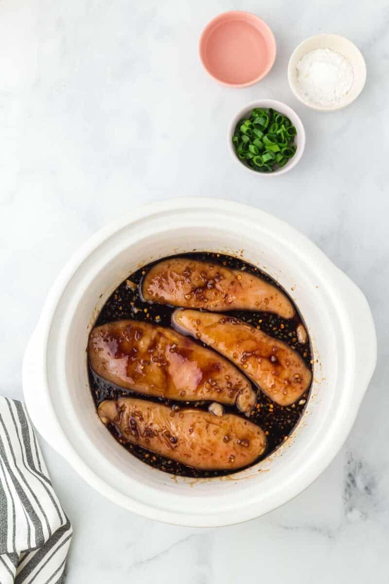 Chicken breasts in dark sauce inside a white slow cooker, with small bowls of sliced green onions, cornstarch, and a pink bowl nearby on a marble surface.