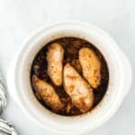 Four cooked chicken breasts sit in a dark sauce inside a round white slow cooker on a light marble surface, next to a striped cloth.