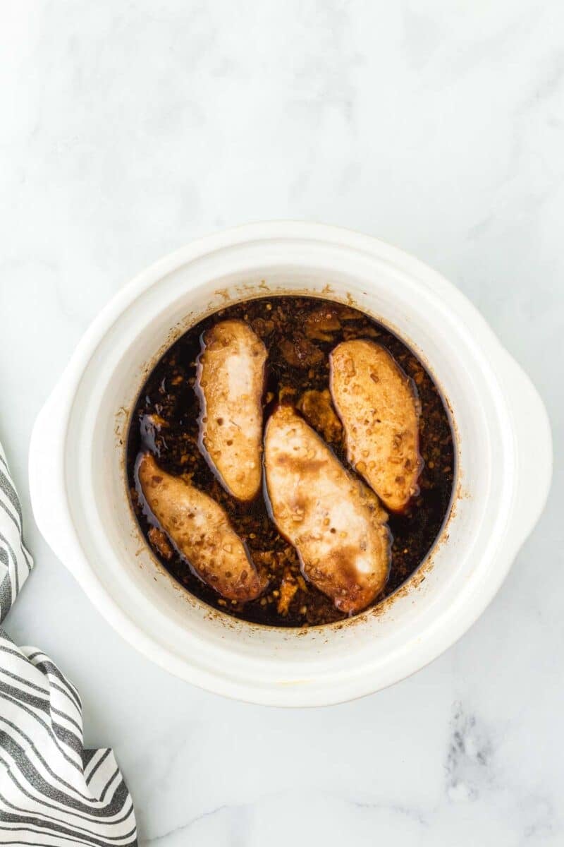 Four cooked chicken breasts sit in a dark sauce inside a round white slow cooker on a light marble surface, next to a striped cloth.
