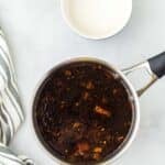 A saucepan with a dark sauce mixture and a bowl of milk on a light surface, next to a striped kitchen towel.