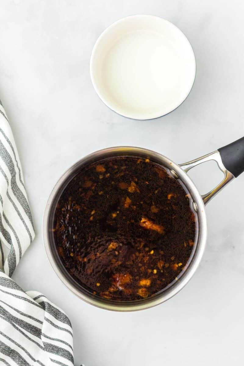 A saucepan with a dark sauce mixture and a bowl of milk on a light surface, next to a striped kitchen towel.