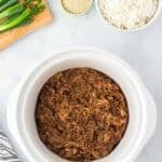 A slow cooker with shredded honey garlic chicken, a bowl of cooked white rice, sliced green onions on a cutting board, and a small bowl of sesame seeds on a marble countertop.