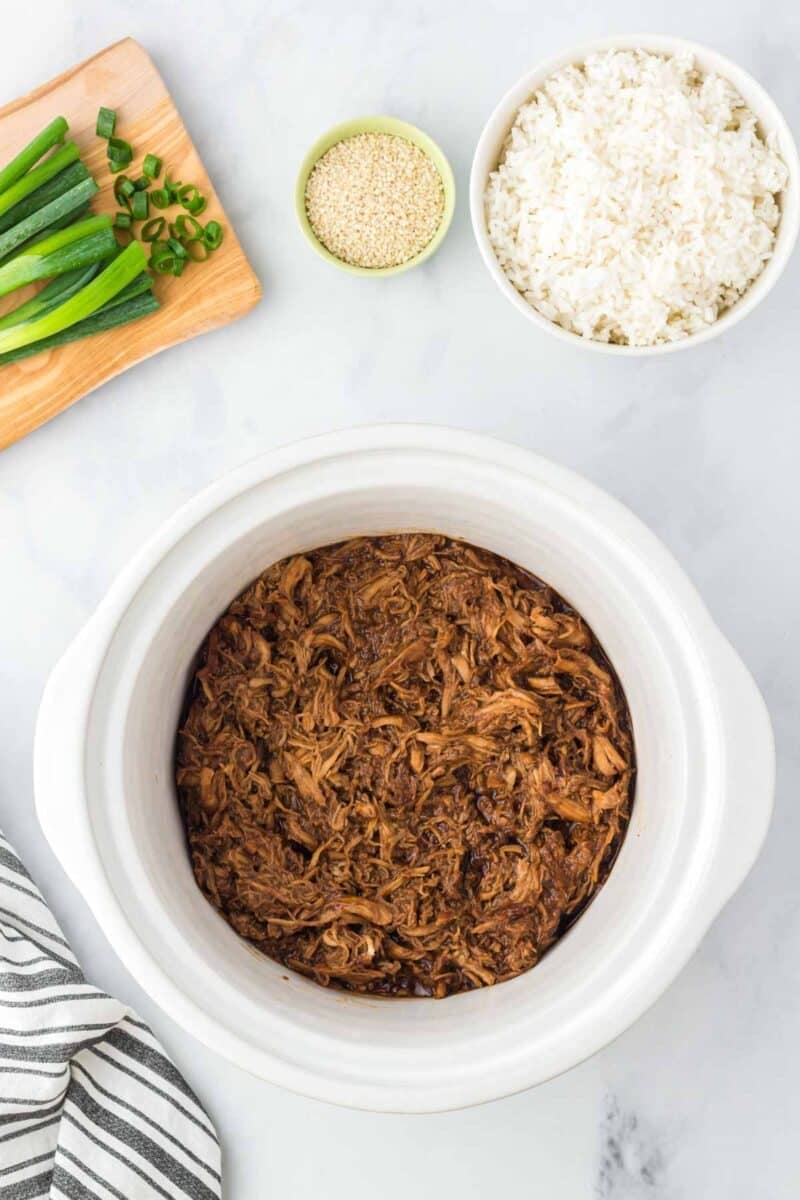 A slow cooker with shredded honey garlic chicken, a bowl of cooked white rice, sliced green onions on a cutting board, and a small bowl of sesame seeds on a marble countertop.
