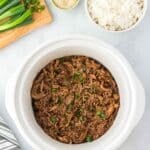Shredded slow cooker honey garlic chicken in a white slow cooker, garnished with green onions, next to a bowl of rice, sesame seeds, and sliced green onions on a wooden board.