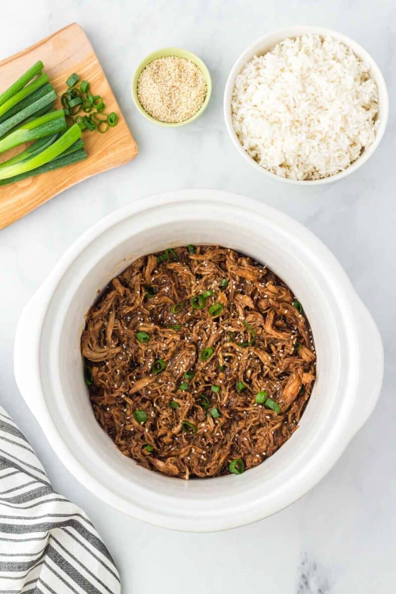 Shredded slow cooker honey garlic chicken in a white slow cooker, garnished with green onions, next to a bowl of rice, sesame seeds, and sliced green onions on a wooden board.