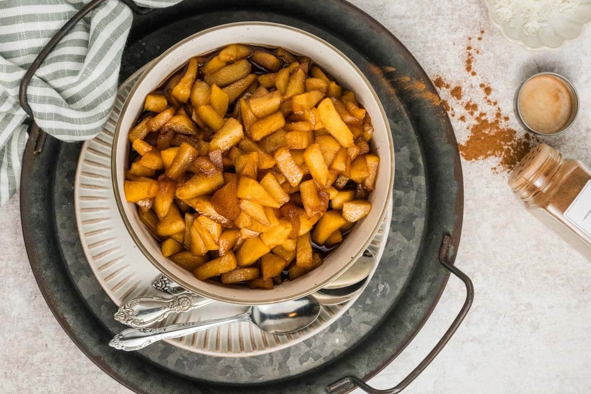 A bowl of cooked cinnamon apples sits on stacked plates with a striped towel, spoons, and a spilled jar of cinnamon nearby on a light surface.