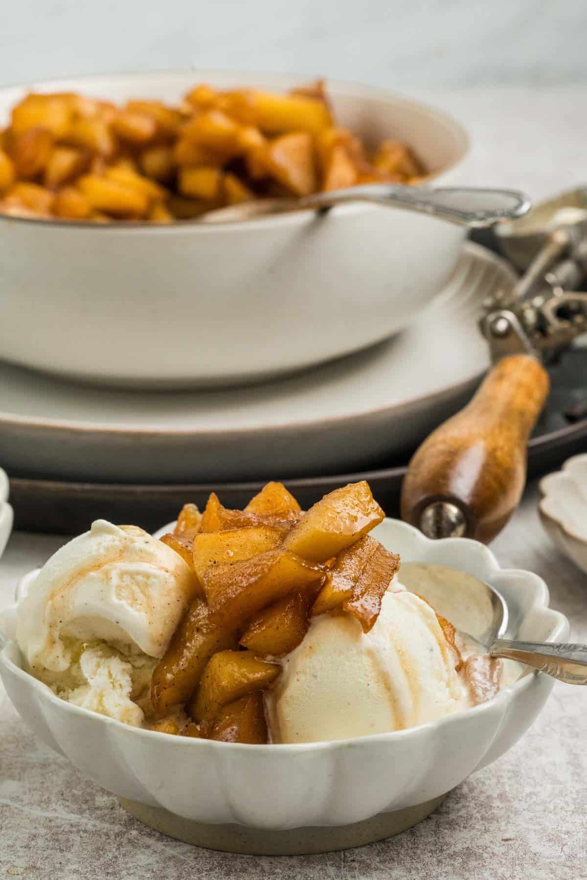 A bowl of vanilla ice cream topped with cooked cinnamon apples, set in front of a bowl filled with more cinnamon apples and a metal ice cream scoop.